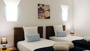 A hotel room with two beds, each with white bedding and patterned pillows. There are wall-mounted lamps and a framed picture above the beds. - Lotus Villa, Praia da Ora - Albufeira (Albufeira)