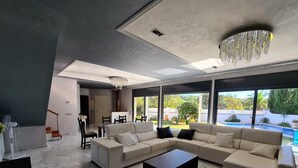 Una sala moderna con un sofá seccional, una mesa de centro y una araña. Hay un comedor con una mesa y sillas, y se ve una piscina y vegetación al exterior.