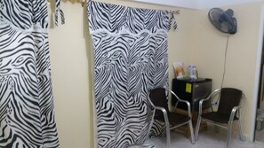Une pièce avec des rideaux à rayures zébrées, une petite table avec des chaises, un ventilateur et un meuble noir sur lequel sont posés divers objets.