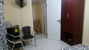 A room with a white door, a red wardrobe, two wicker chairs, a small table with a bottle and a box, and a zebra-patterned rug.