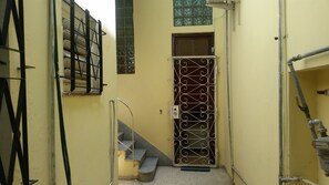 Un couloir étroit avec une grille métallique, un escalier et une conduite d’eau.