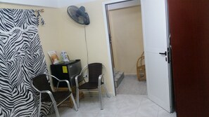 Une pièce avec un rideau à motifs zébrés, un bureau avec un ordinateur et des papiers, un ventilateur et une porte donnant sur une autre pièce.