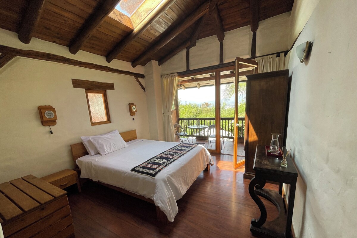 A bedroom with a wooden bed, a nightstand, a dresser, a wooden table with a lamp, and a balcony with chairs.