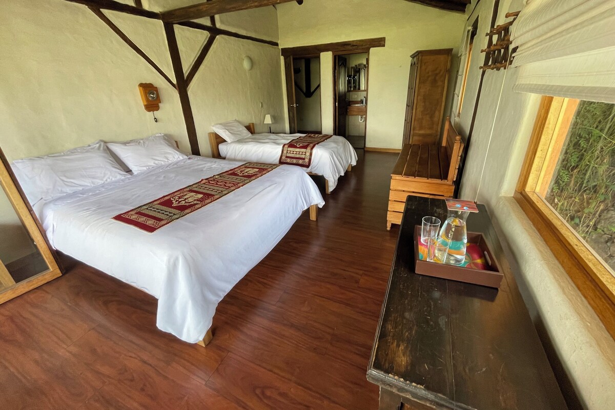 A hotel room with two beds, a wooden bench, a small table with a tray, and a window with a view of greenery.