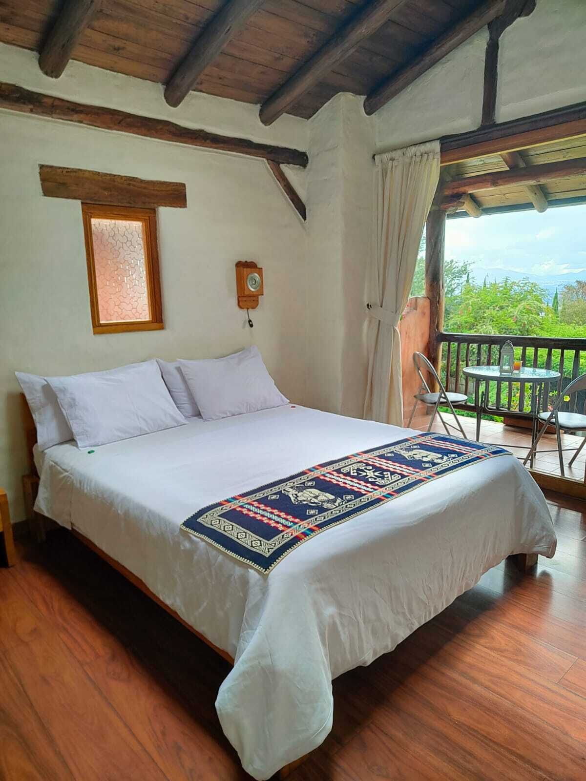 A bedroom with a bed, a wooden ceiling, a window, a wall-mounted phone, and a balcony with chairs.