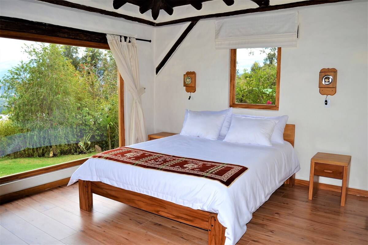Une chambre avec un lit en bois, une literie blanche, un plaid rouge et blanc, une table de chevet en bois, une commode en bois, une fenêtre donnant sur un paysage verdoyant et une horloge murale.