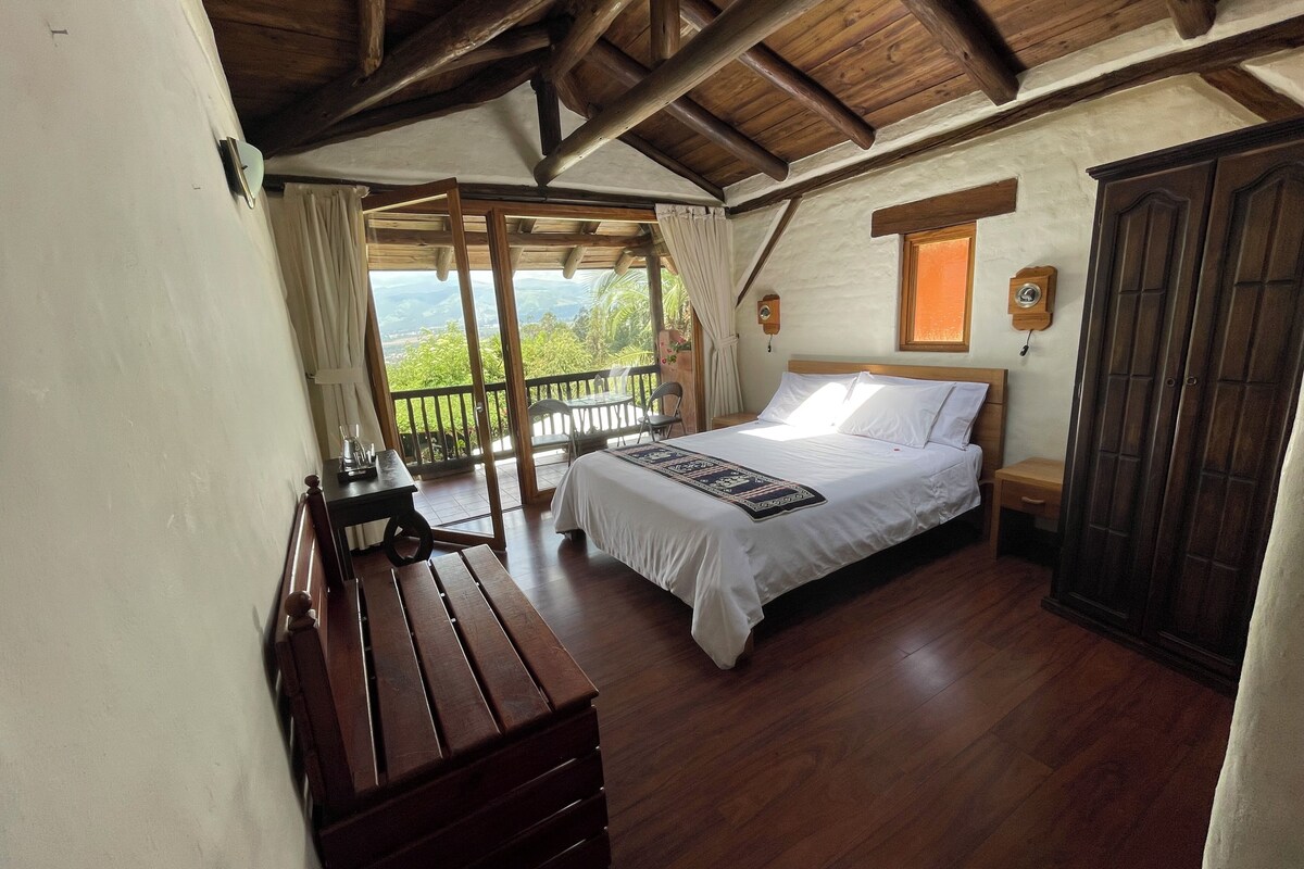 A bedroom with a wooden bed, a wooden nightstand, a wooden bench, and a view of the outdoors.