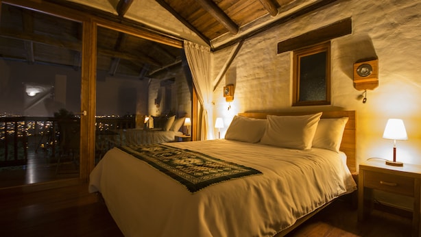 Un dormitorio con una cama grande, vigas de madera en el techo, una mesita de noche con una lámpara y vistas a través de una puerta de cristal.