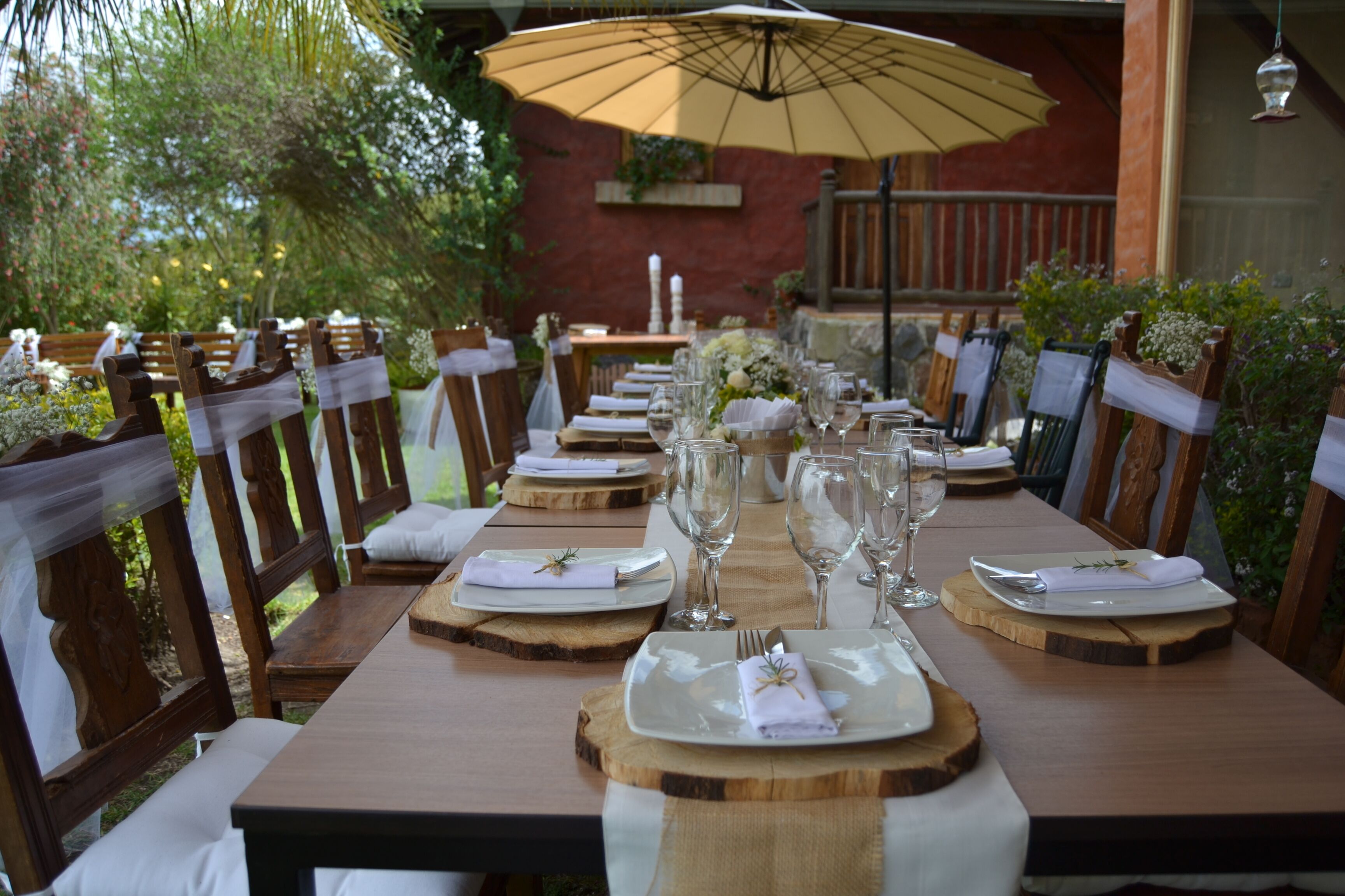 A long wooden dining table set with white plates, silverware, and wine glasses, surrounded by wooden chairs with white cushions.