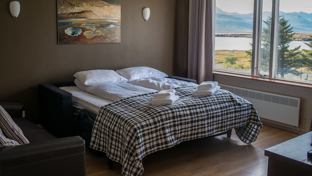 A hotel room with a bed, a painting on the wall, and a window with a view of mountains and a lake. - The View Apartments (Fjardabyggd)