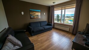 A living room with a dark sofa, a wooden floor, a painting on the wall, and a large window with a view of a landscape. - The View Apartments (Fjardabyggd)