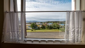 A window with lace curtains, a view of a town with houses and mountains in the distance. - The View Apartments (Fjardabyggd)