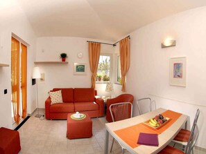 Un espacio habitable compacto con sofá, una mesa de comedor con sillas, una mesita con lámpara y una ventana con cortinas.