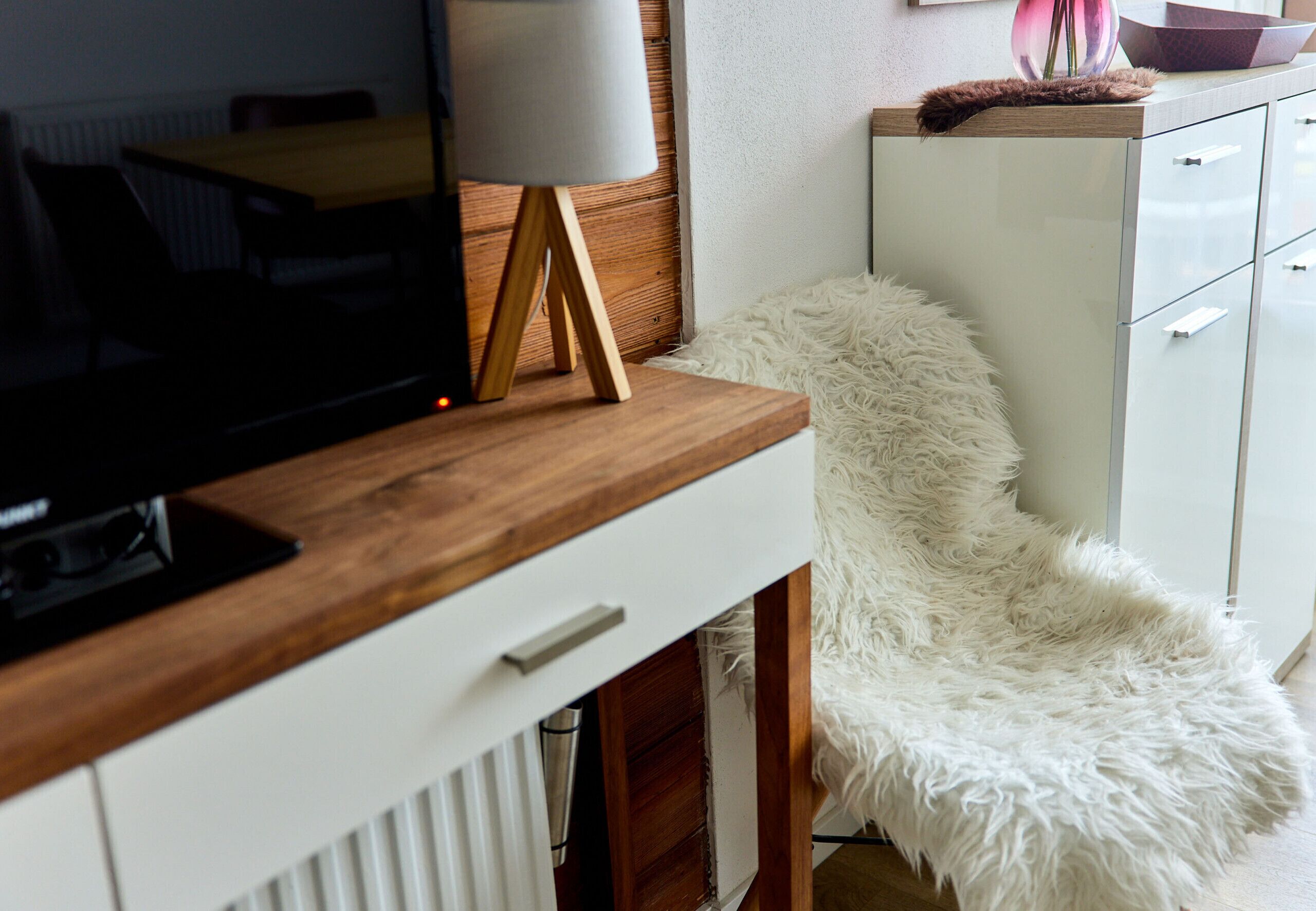 A modern room with a wooden desk, a fluffy white chair, a white cabinet, and a television.