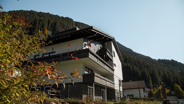 A two-story building with a balcony on the upper floor, surrounded by trees and a forested mountain in the background. - 1105 Veilchen (Gaschurn)