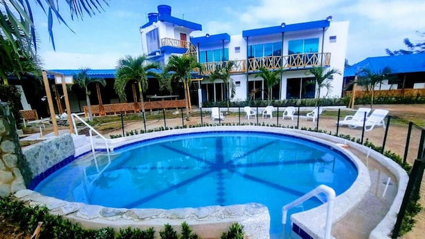 A swimming pool with a stone border, surrounded by lounge chairs and a building with blue and white exterior. - Cabañas Vacacionales En Palomino Guajira (Dibulla)