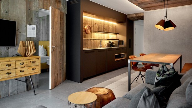 A modern kitchen with wooden accents, a dining area with a wooden table and chairs, and a living area with a sofa and ottoman.