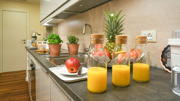 Una cocina moderna con un lavamanos de acero inoxidable, un frutero con manzanas y una piña, y tres botellas de vidrio con jugo de naranja.