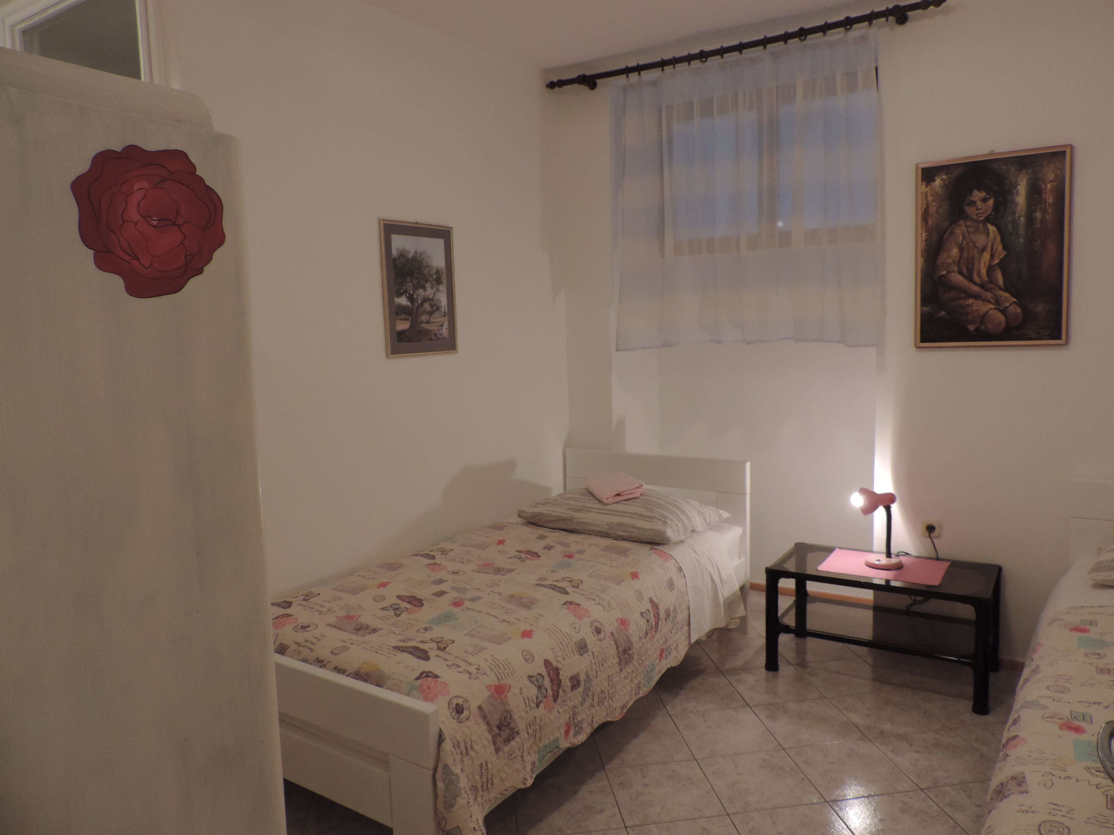 A small, well-lit bedroom with a single bed, a nightstand, a window with sheer curtains, and a wall-mounted picture.