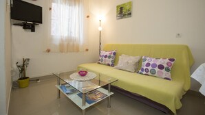 A living room with a yellow sofa, a glass coffee table, a potted plant, a wall-mounted TV, and a window with sheer curtains. - Apartment Pera (Split)
