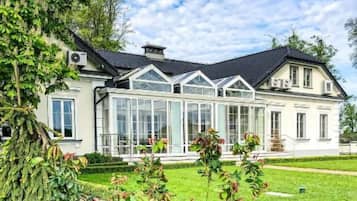 Una casa grande de dos pisos con un invernadero, rodeada de un jardín cuidado y un cielo despejado.