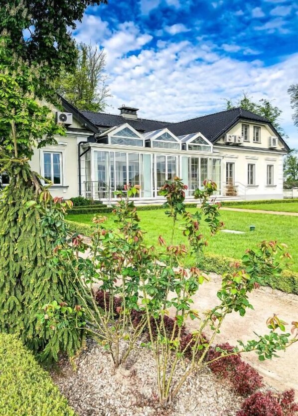 A large, two-story house with a conservatory, surrounded by a well-maintained garden and a clear sky.