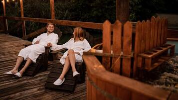 Two individuals seated on wooden loungers on a wooden deck with string lights overhead.