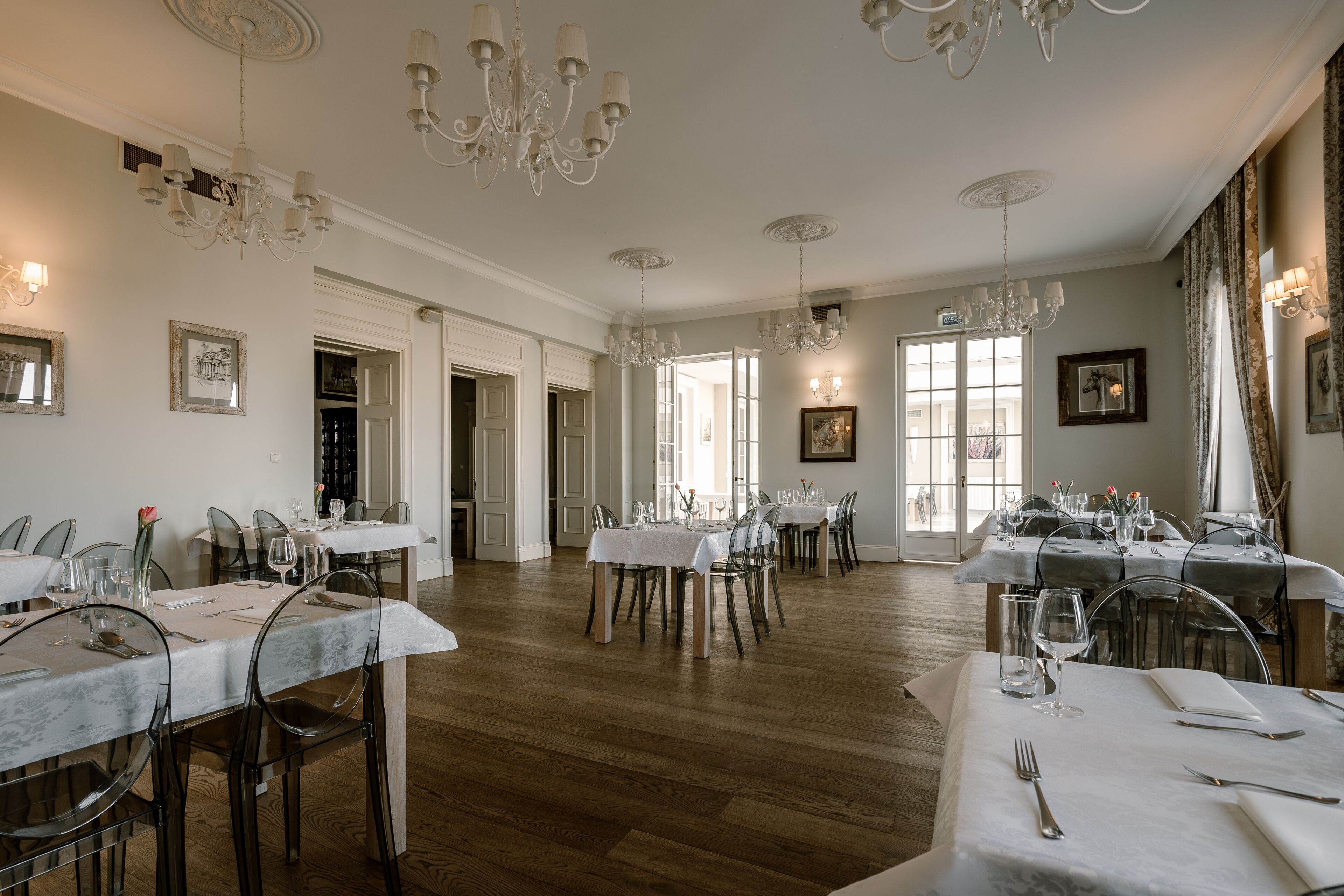 A spacious dining area with multiple tables set for dining, elegant chandeliers, and framed artwork on the walls.
