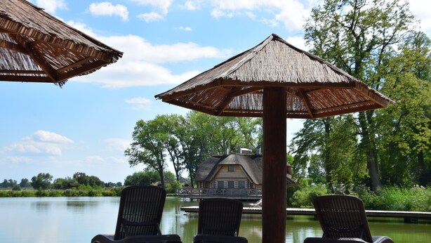 A beach area with two lounge chairs under a thatched umbrella, a wooden post, and a view of a house across a body of water. - Hotel Przy Dworze (Lesna Podlaska)
