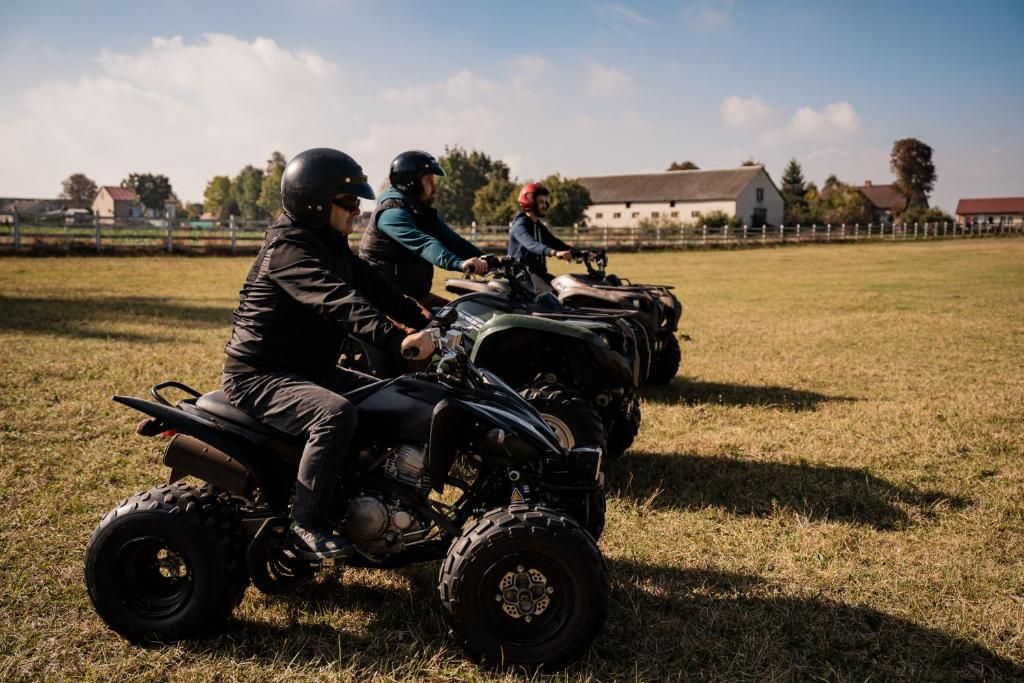 Three individuals on all-terrain vehicles in a grassy field with buildings in the background.