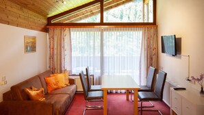 Une pièce avec un plafond en bois, un canapé avec des coussins orange, une table à manger avec des chaises, un téléviseur fixé au mur et une fenêtre avec des rideaux.