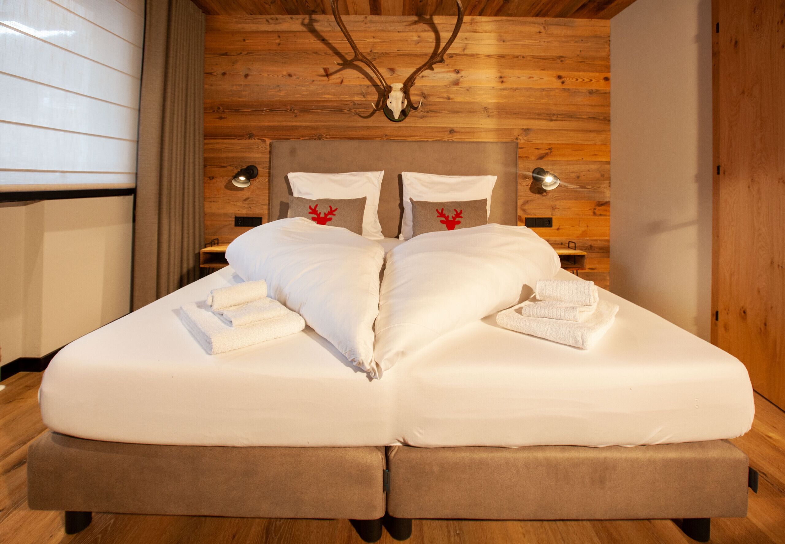 Una cama doble con sábanas blancas y dos almohadas, techo de madera con decoración de cráneo de ciervo, y dos lámparas de noche.