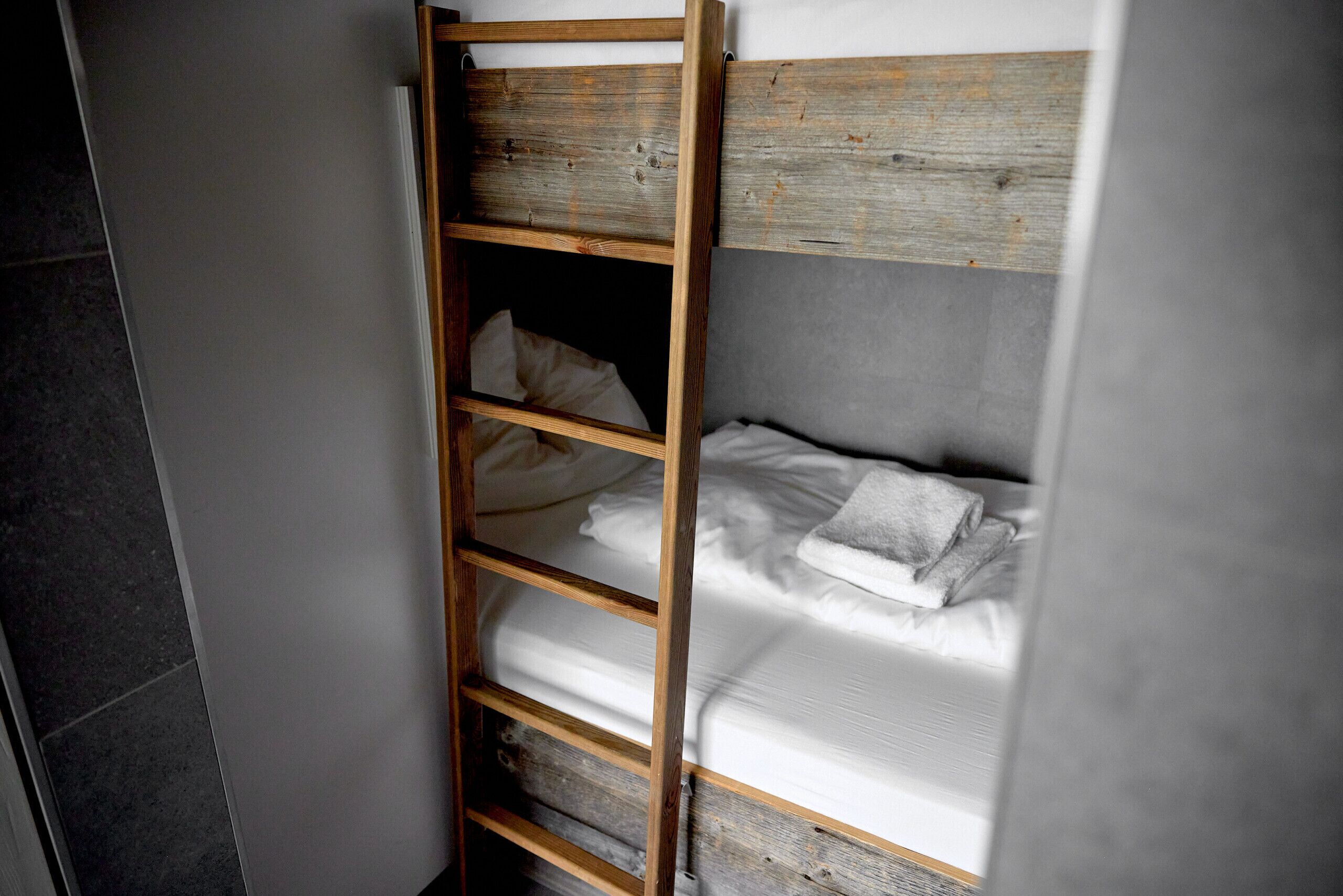 Een stapelbed met houten hoofdeinde en een ladder, wit beddengoed en een grijze muur.