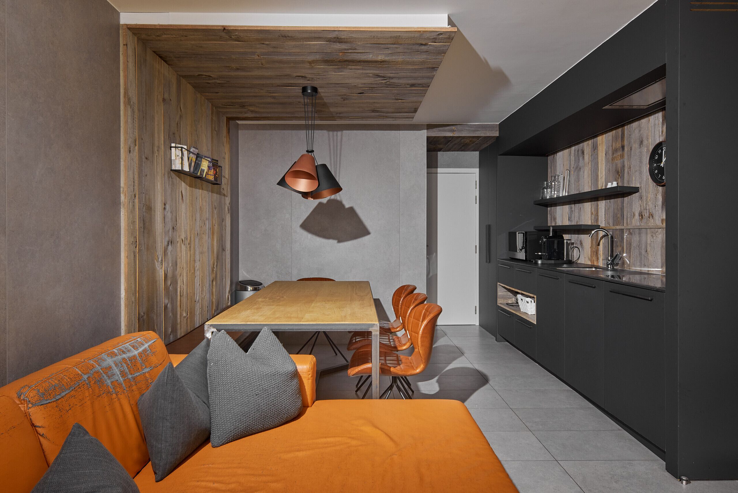 A modern kitchen with dark cabinetry, a wooden ceiling, and a dining area with a wooden table and orange chairs.