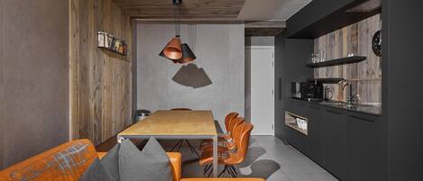 A modern kitchen with dark cabinetry, a wooden ceiling, and a dining area with a wooden table and orange chairs.