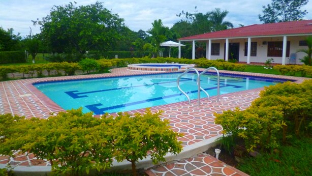 A rectangular swimming pool with a tiled deck, surrounded by well-maintained greenery and a building with a red roof in the background. - Finca Vacacional Villavicencio (Villavicencio)