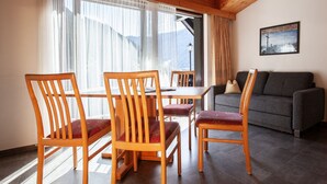 Une pièce comprenant une table et des chaises en bois, un canapé gris et une grande fenêtre ornée de rideaux légers.