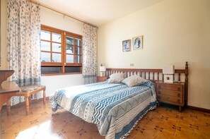 Une chambre à coucher avec un lit, une table de chevet en bois, une chaise, une fenêtre avec des rideaux et un mur orné de deux tableaux encadrés.
