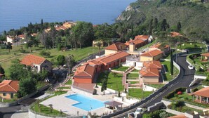 Un complejo turístico en la costa con edificios de techo de teja terracota, una piscina central y un camino sinuoso.