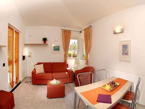 Un espacio habitable compacto con sofá, una mesa de comedor con sillas, una mesita con lámpara y una ventana con cortinas.
