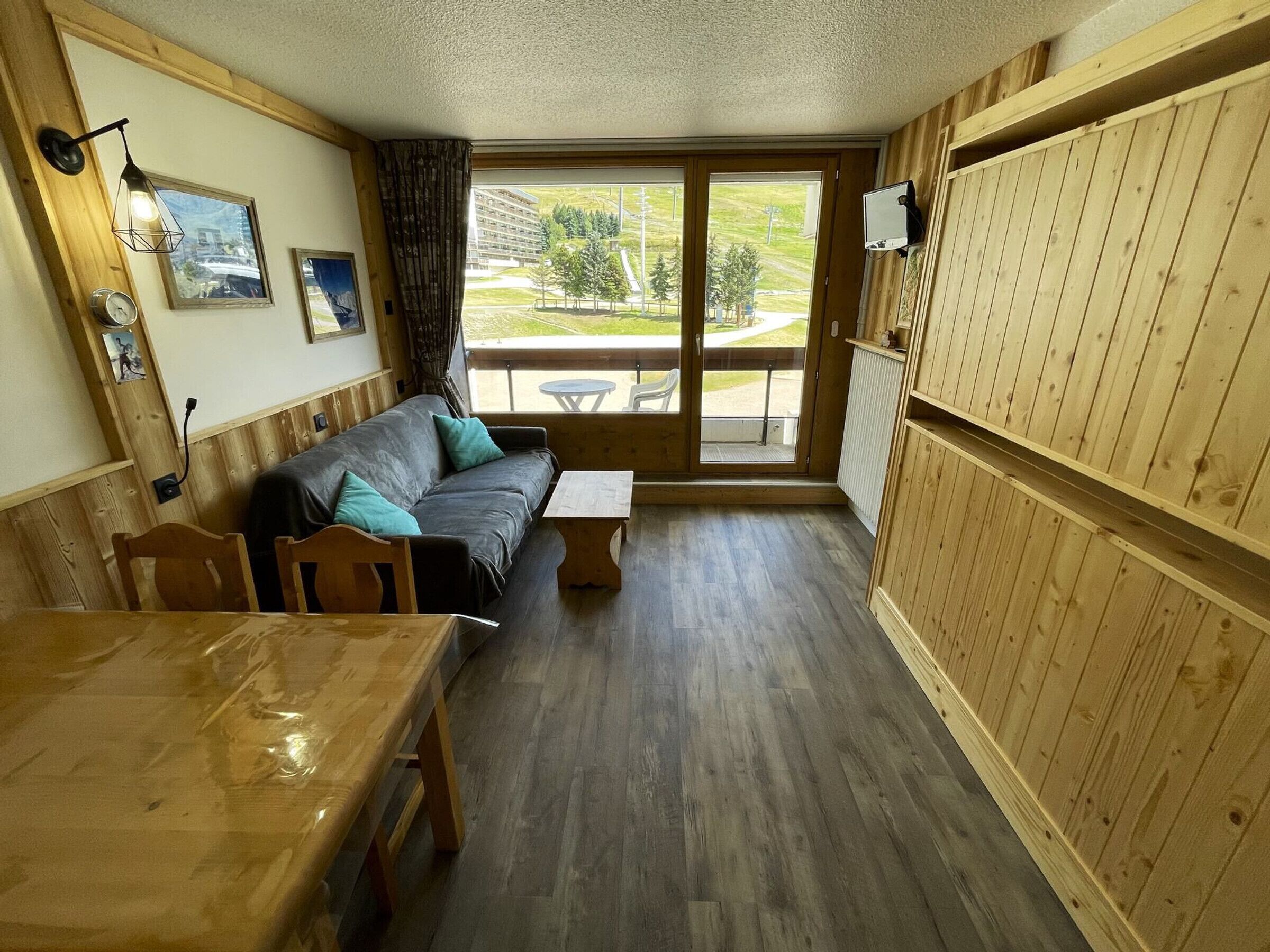 A cozy room with a wooden floor, a sofa with teal cushions, a wooden table, and a large window offering a view of the outdoors.