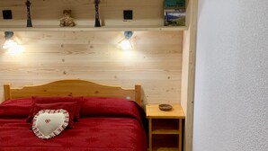 A bedroom with a wooden headboard, a red bedspread, a heart-shaped pillow, a small wooden nightstand, and a shelf with decorative items and a book. - 3-room apartment for 4/6 people in Valfréjus with balcony (Valfréjus)