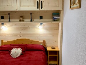 A bedroom with a wooden headboard, a red bedspread, a heart-shaped pillow, a small wooden nightstand, and a shelf with decorative items and a book. - 3-room apartment for 4/6 people in Valfréjus with balcony (Valfréjus)