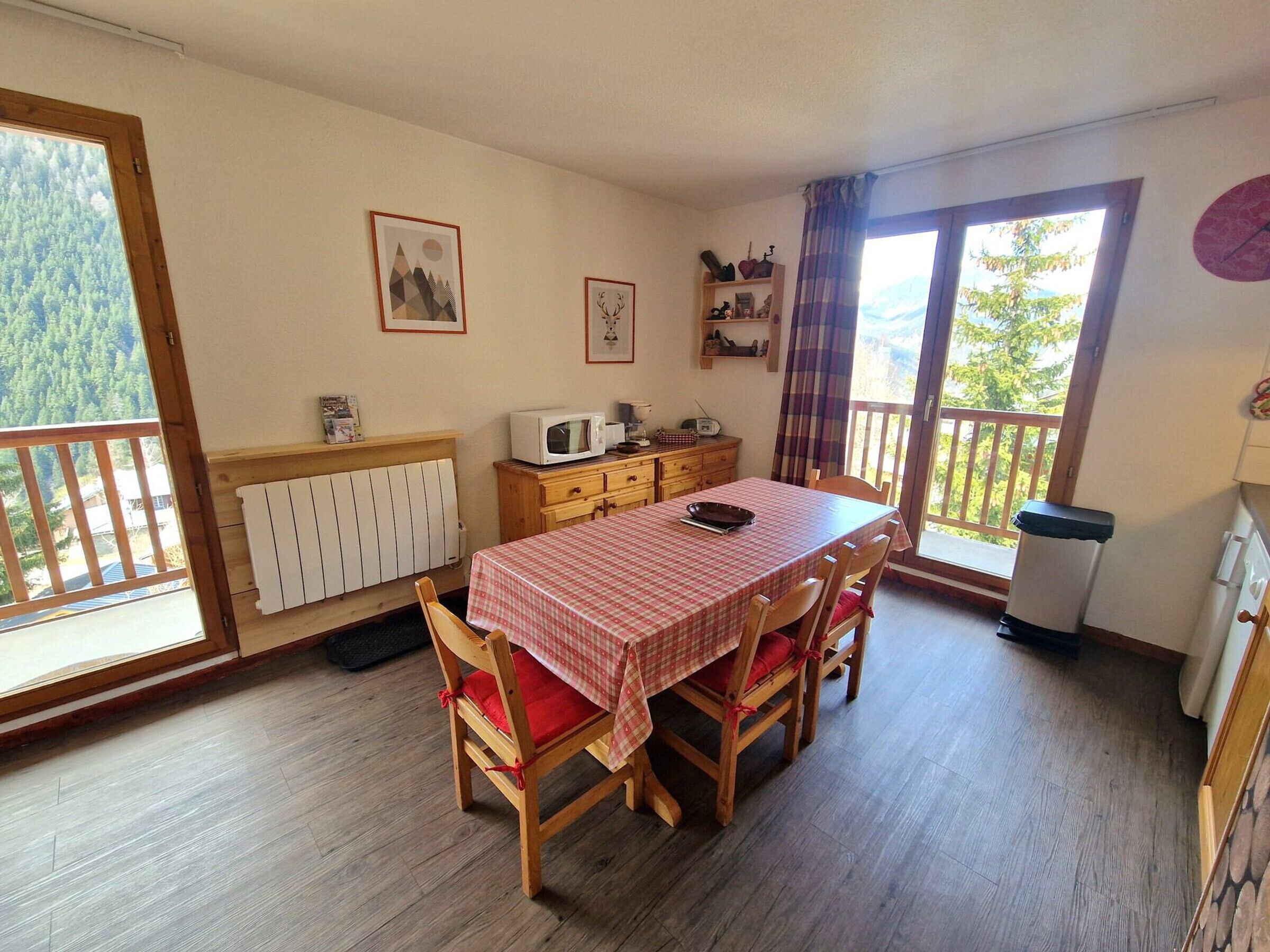 Un comedor con una mesa cubierta por un mantel a cuadros, sillas de madera con cojines rojos, un radiador blanco, un mueble de madera con un microondas y una ventana con vistas a árboles.