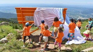 Un grupo de niños con camisetas anaranjadas interactuando con un lienzo grande blanco y azul en una ladera.