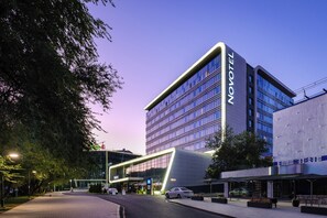 A modern hotel building with a prominent 'NOVOTEL' sign, a covered outdoor area with seating, and a parking lot. - Novotel Almaty City Center (Almaty)