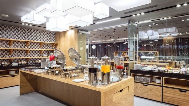 A modern breakfast bar with glass-fronted display cases, wooden shelves, and a variety of breakfast items. - Novotel Almaty City Center (Almaty)