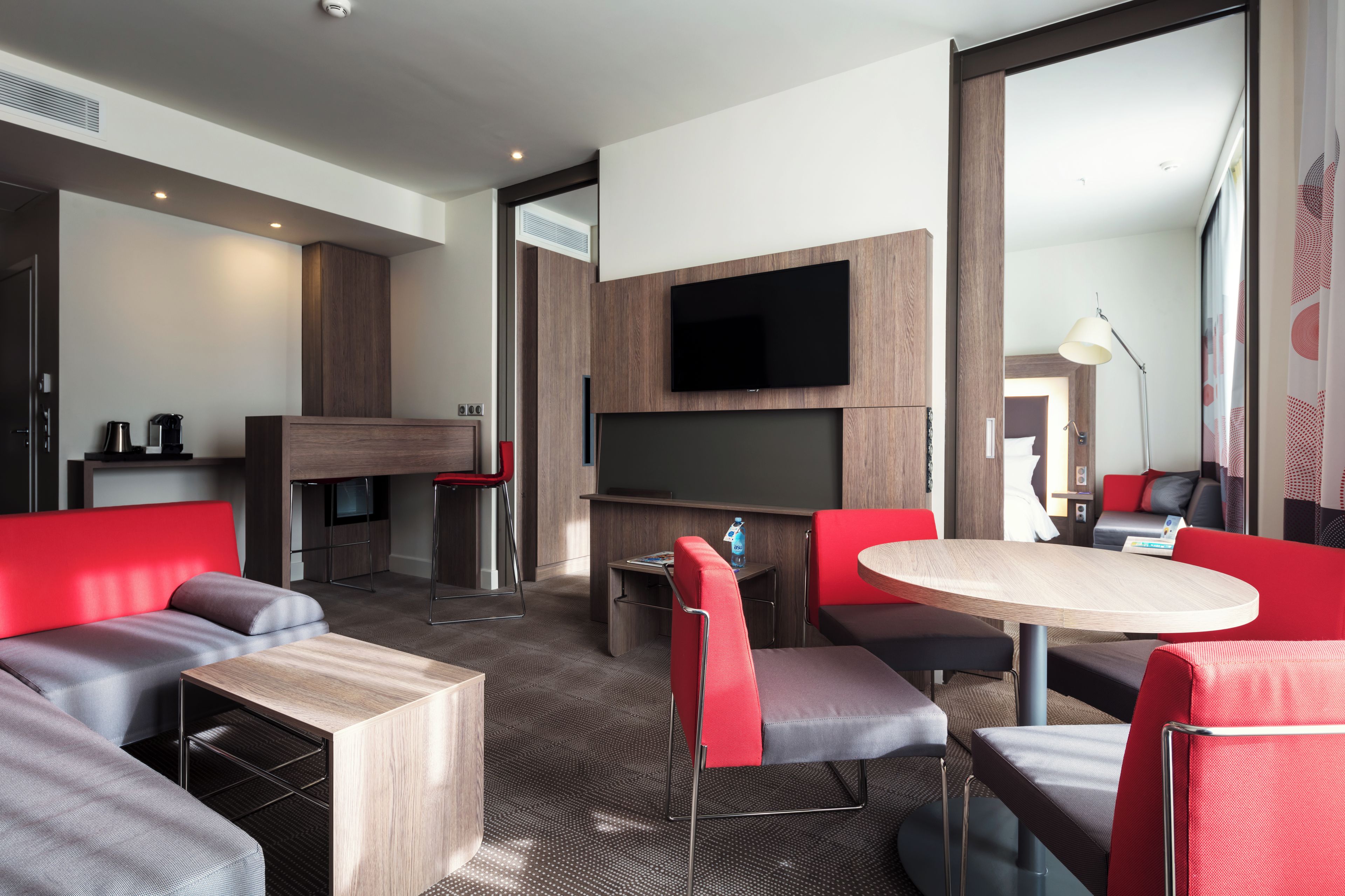 A modern hotel room with a flat-screen TV, a dining area with a round table, and a seating area with red and grey upholstered furniture.