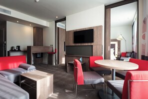 A modern hotel room with a flat-screen TV, a dining area with a round table, and a seating area with red and grey upholstered furniture. - Novotel Almaty City Center (Almaty)