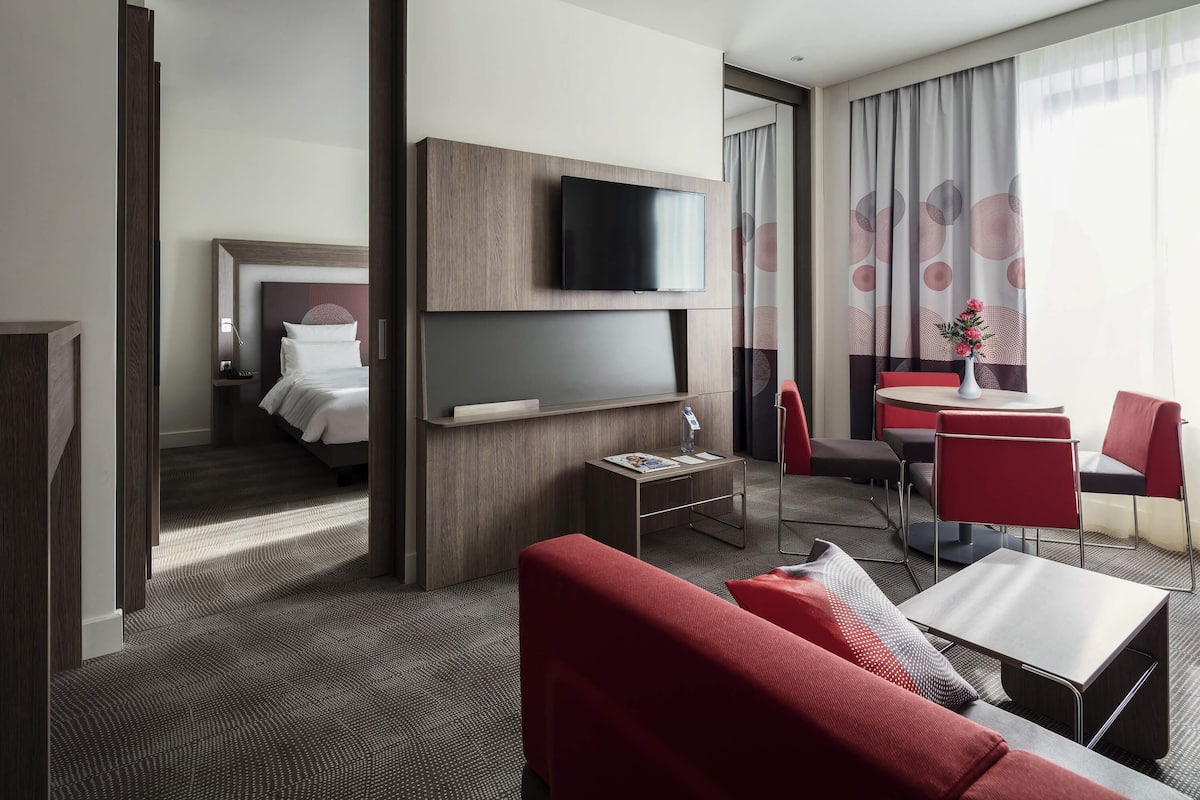 Une chambre d’hôtel comprenant un lit, un téléviseur fixé au mur, un bureau avec une chaise, un canapé rouge et un coin repas avec une table et des chaises.